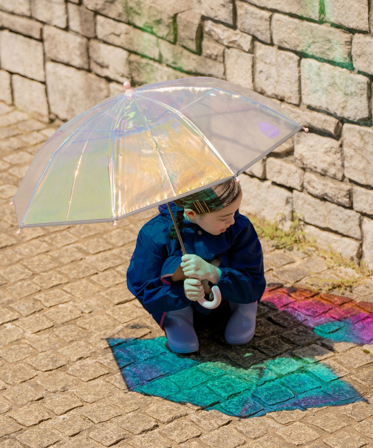 kid holding a transparent umbrella on a stone pavement with a rainbow-colored reflection on the floow
